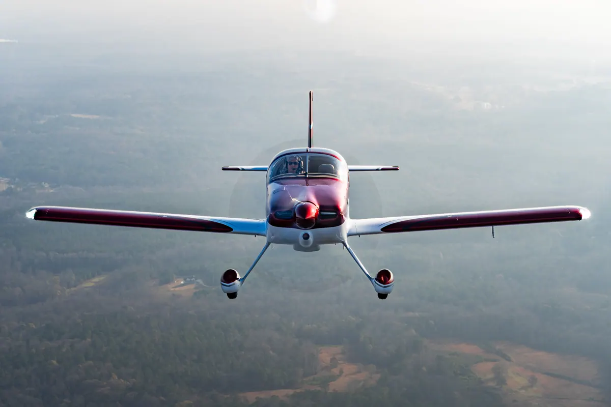 The red-and-white low-wing homebuilt flies straight at the camera, wings level, over hazy woods