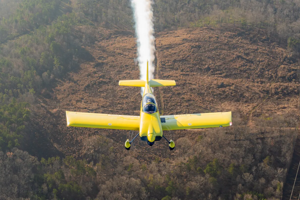 Closer view of the yellow low-wing homebuilt trailing smoke over cleared land and woods