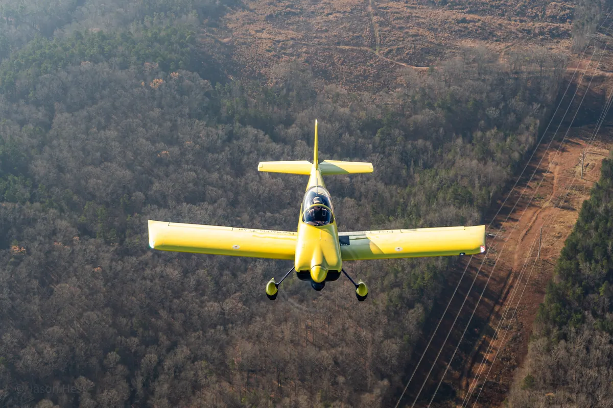 The yellow low-wing homebuilt flies head-on above winter woods and a power-line cut
