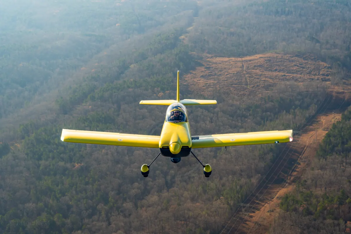 The yellow low-wing homebuilt flies head-on over winter woods, pilot visible under the canopy