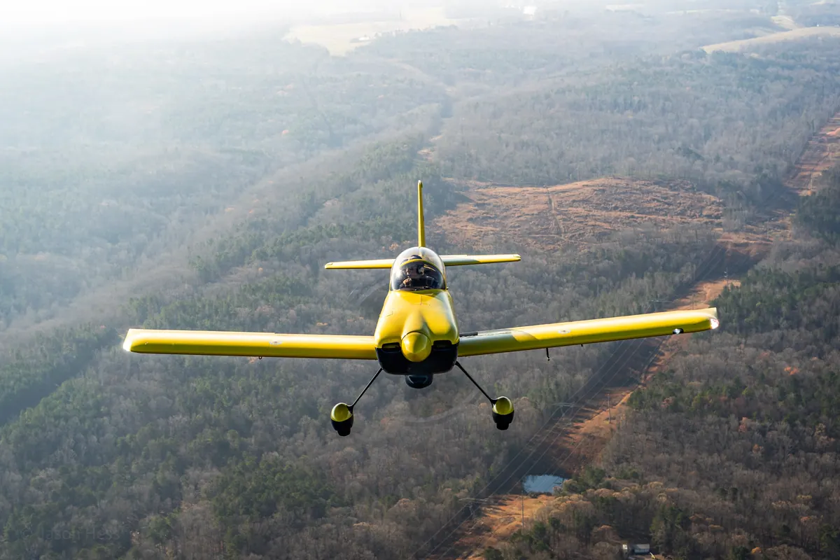 The yellow low-wing homebuilt flies head-on over wooded ridges in soft winter haze