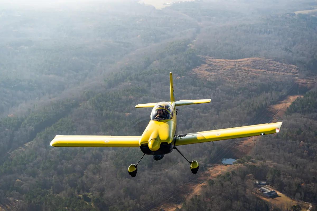 The yellow low-wing homebuilt banks slightly toward the camera above wooded hills