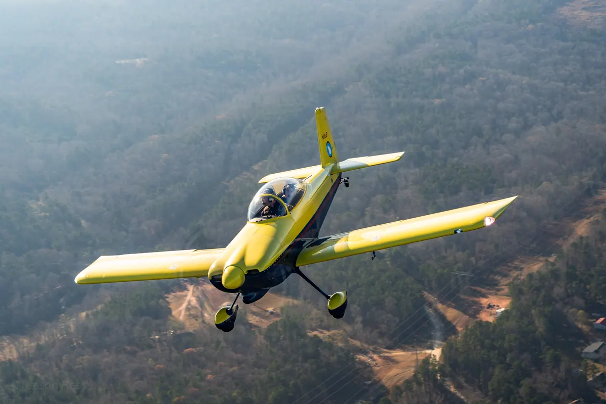 The yellow low-wing homebuilt banks in toward the camera, sunlight on its wings, over wooded hills