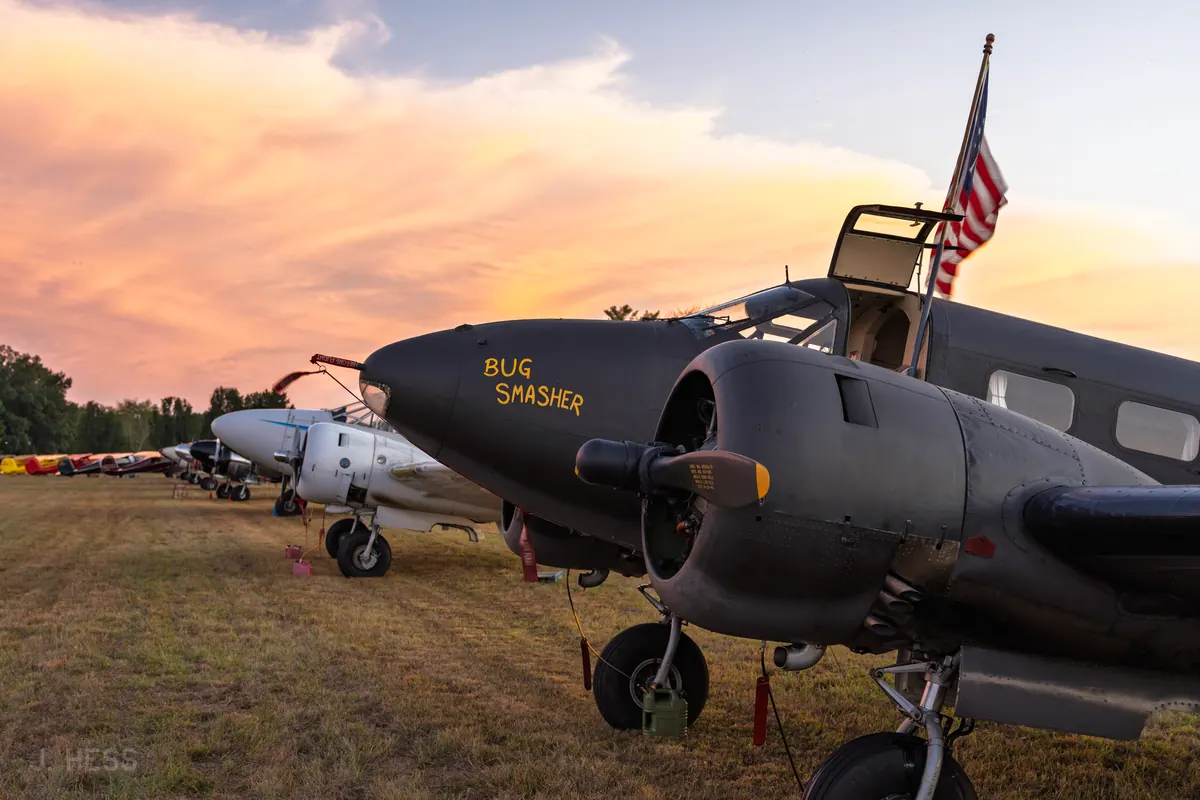 Three-quarter view of the olive-drab 'Bug Smasher' Beech 18 at dusk, with a long row of vintage aircraft fading into the background