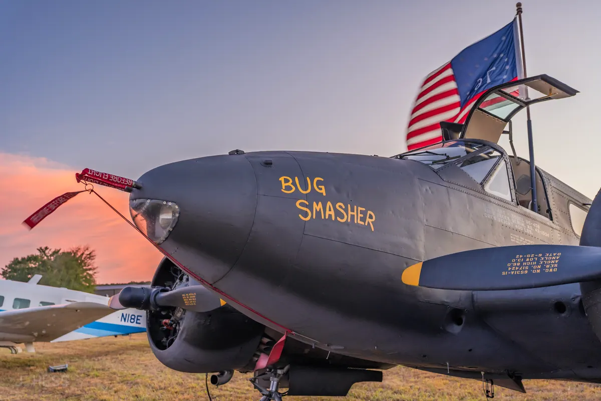 Close view of the olive-drab nose of the Beech 18 'Bug Smasher', its name painted in yellow, with a Bicentennial flag flying above the cockpit at sunset