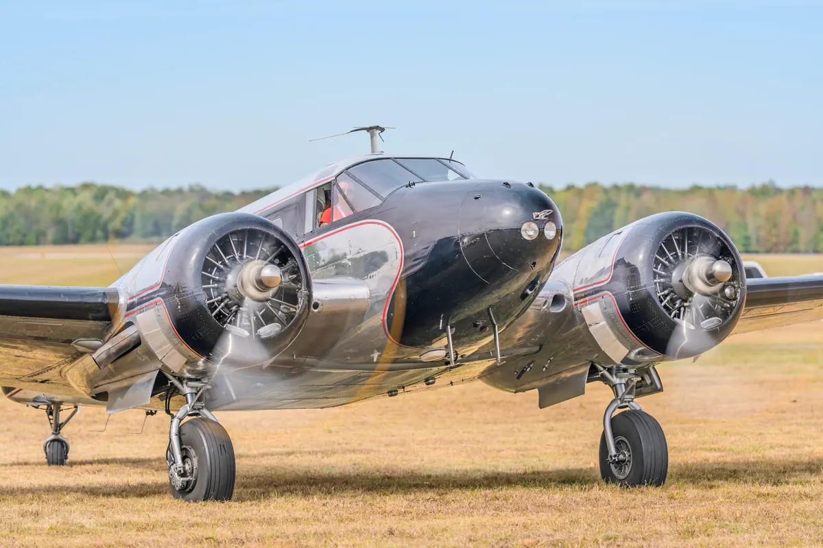 Close three-quarter view of a polished Beech 18 with a black nose and red pinstripes, propellers turning, on a grass field under a clear blue sky