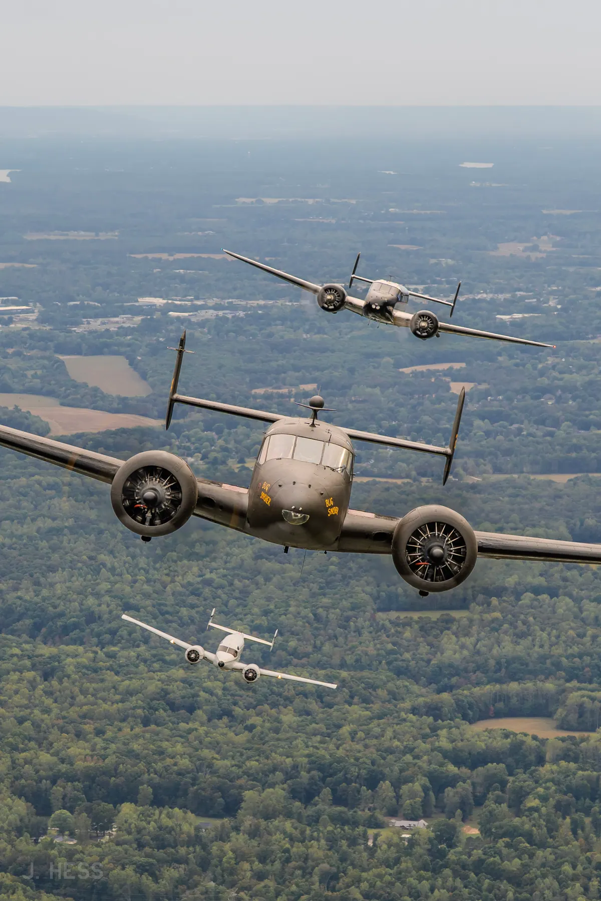 An olive-drab Beech 18 leads a silver and a white Twin Beech in a stacked formation over forested hills