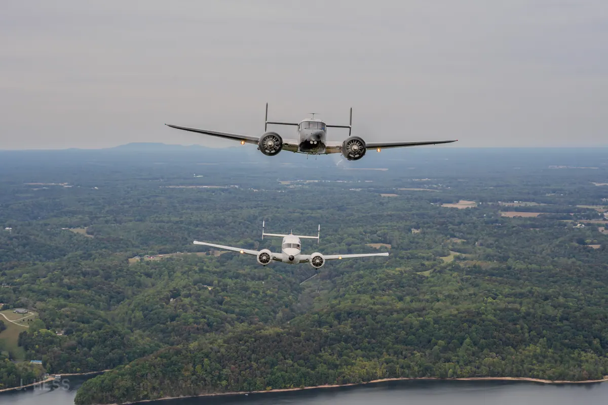 A polished Beech 18 flies straight toward the camera, landing lights on, with a white Twin Beech lower and behind, over wooded hills and the edge of a lake