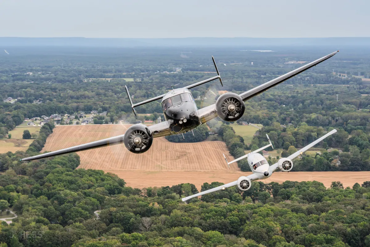 A polished Beech 18 banks toward the camera with a white Twin Beech banking alongside, over a harvested field and woods