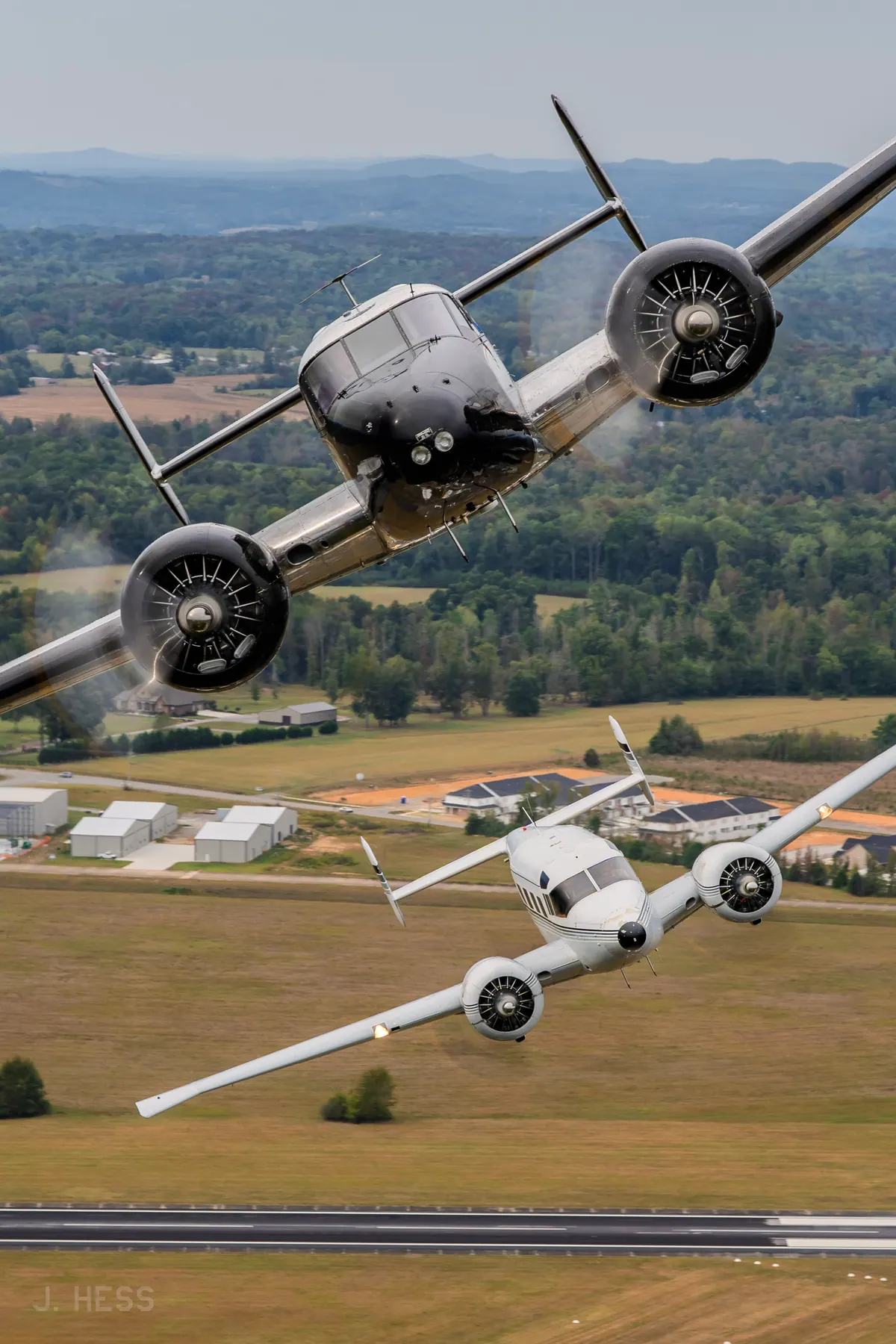 A polished Twin Beech banks steeply toward the camera with a white Twin Beech close below it, over an airfield with hangars and a runway