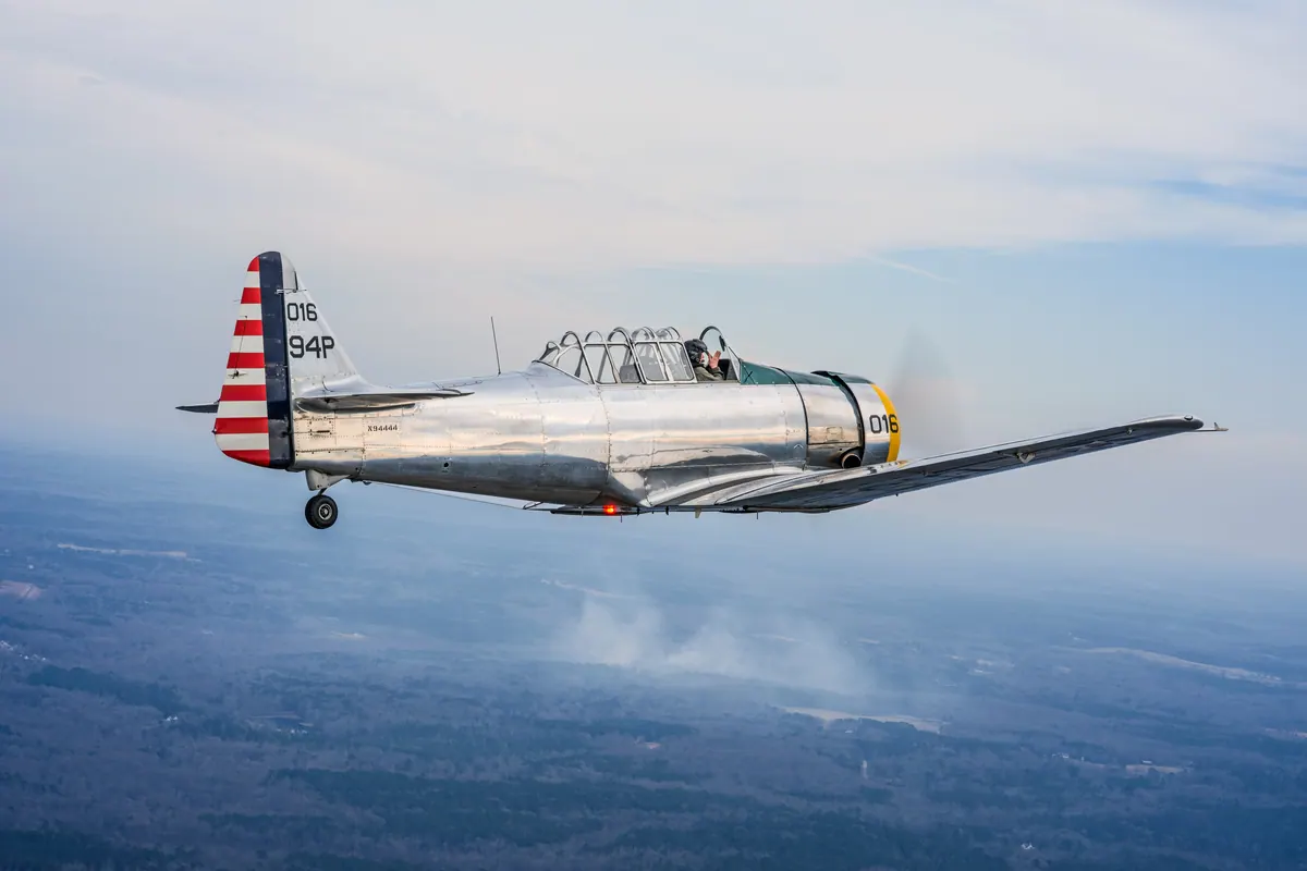 Side view of the polished silver trainer '016', red-and-white striped rudder, flying over a blue haze with a puff of cloud below