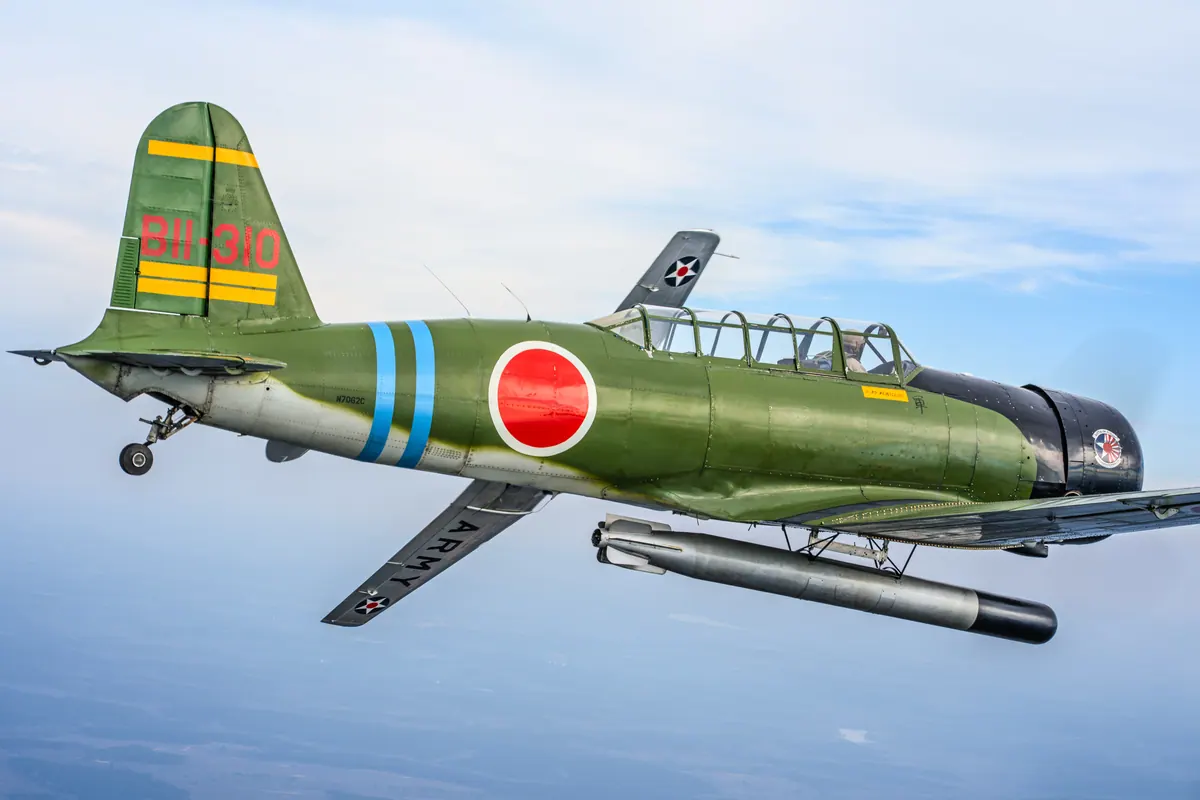 Close side view of the green 'Kate' torpedo-bomber replica, red roundel and blue fuselage bands, a dummy torpedo slung underneath, against a blue sky