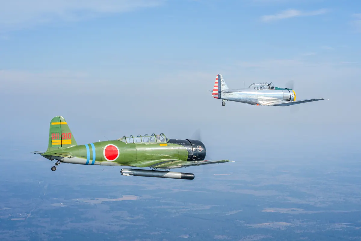 The green 'Kate' torpedo-bomber replica below and the silver trainer above and behind, over a hazy blue landscape