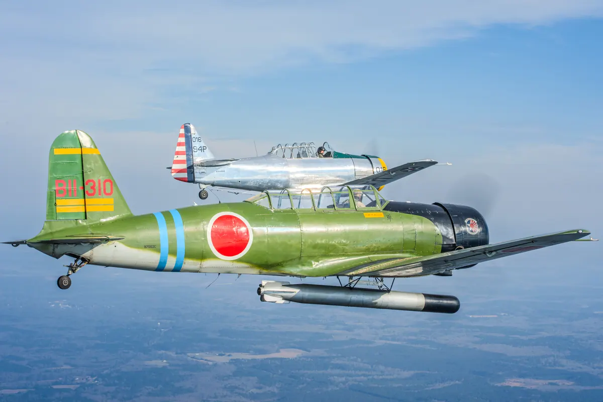 The green 'Kate' torpedo-bomber replica with the silver trainer tucked in just above and behind it, over hazy countryside