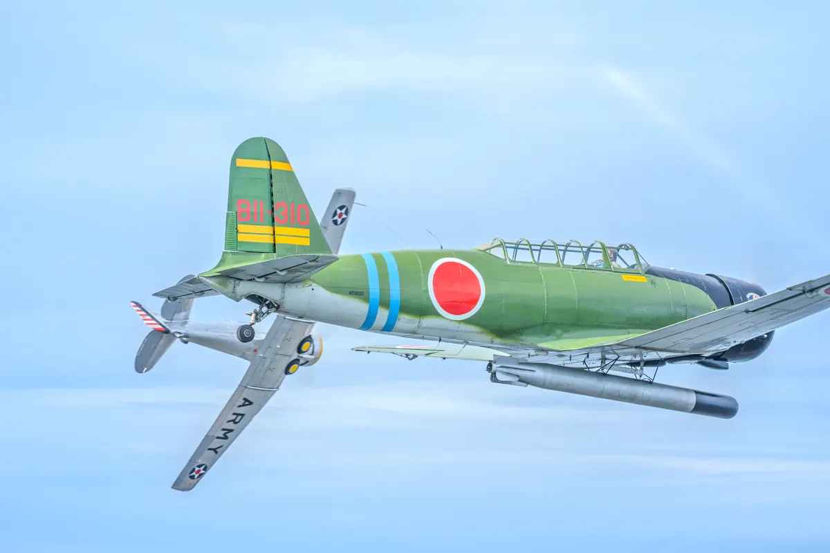 The green 'Kate' torpedo-bomber replica passes in front of the banking silver trainer under a pale blue sky
