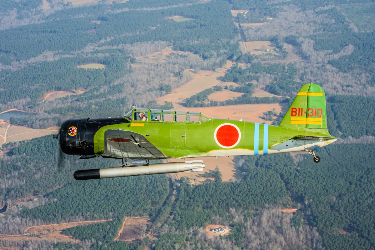 The green 'Kate' torpedo-bomber replica flies over pine forest and cleared fields