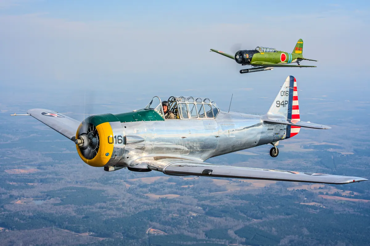 The polished silver trainer '016' in front, the green 'Kate' torpedo-bomber replica higher up and behind, over hazy farmland
