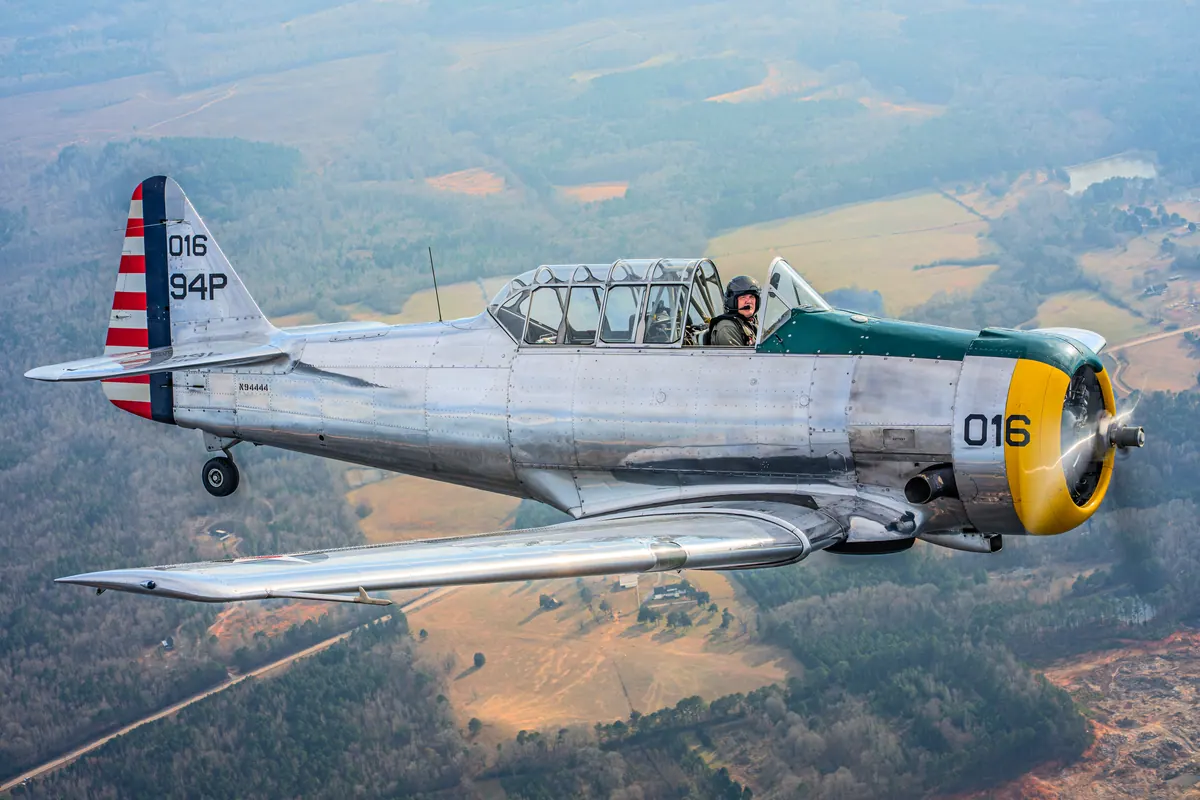 Close side view of the polished silver trainer '016' with its yellow cowl and green anti-glare panel, over farmland