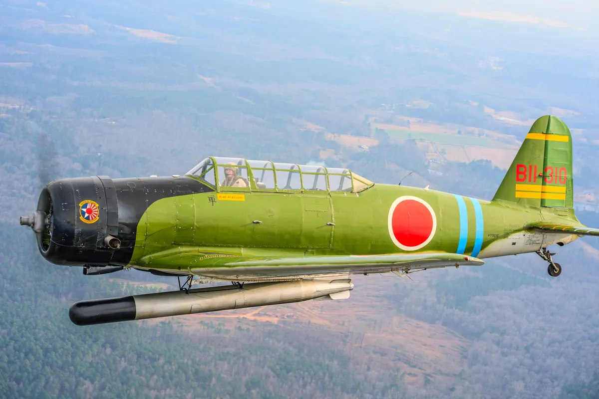 The green 'Kate' torpedo-bomber replica flies left above forest and cleared land, its torpedo slung underneath