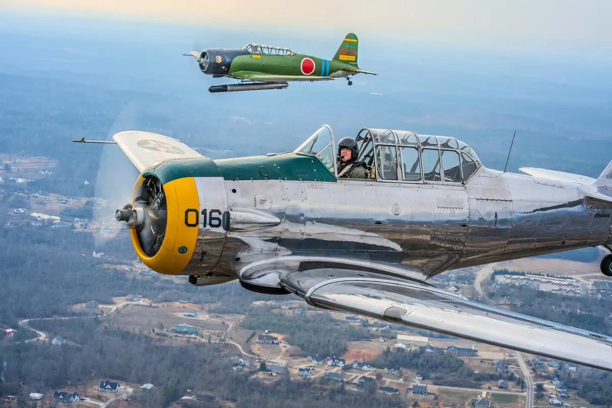 The polished silver trainer '016' flies over a town in warm late light, with the green 'Kate' torpedo-bomber replica high above